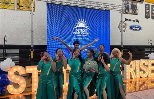 **Alt Text:**   A group of eight Henry Ford High School dancers pose joyfully in matching emerald green costumes after their 